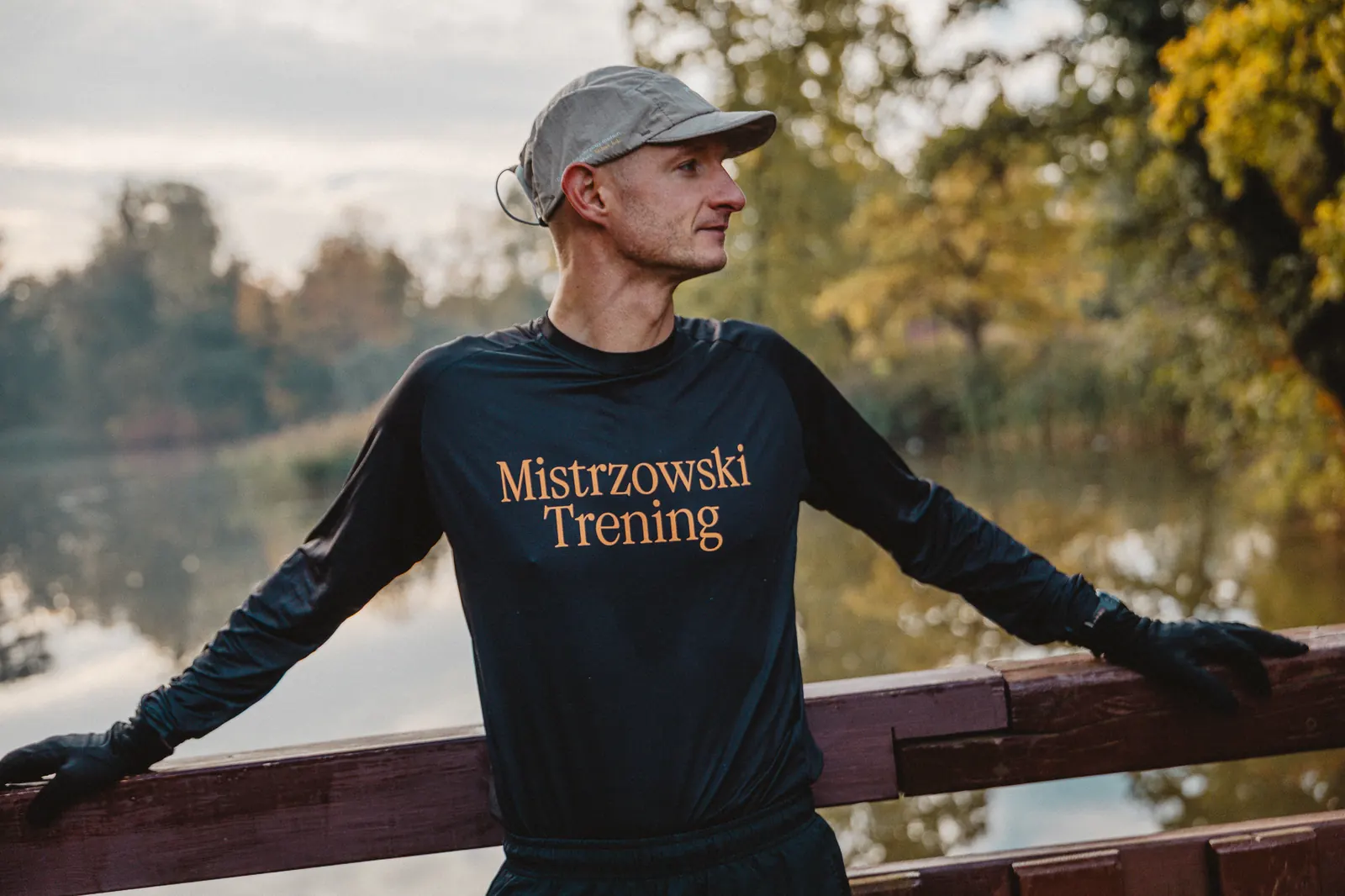 Marcin w koszulce Mistrzowski Trening opiera się o drewnianą barierkę nad stawem